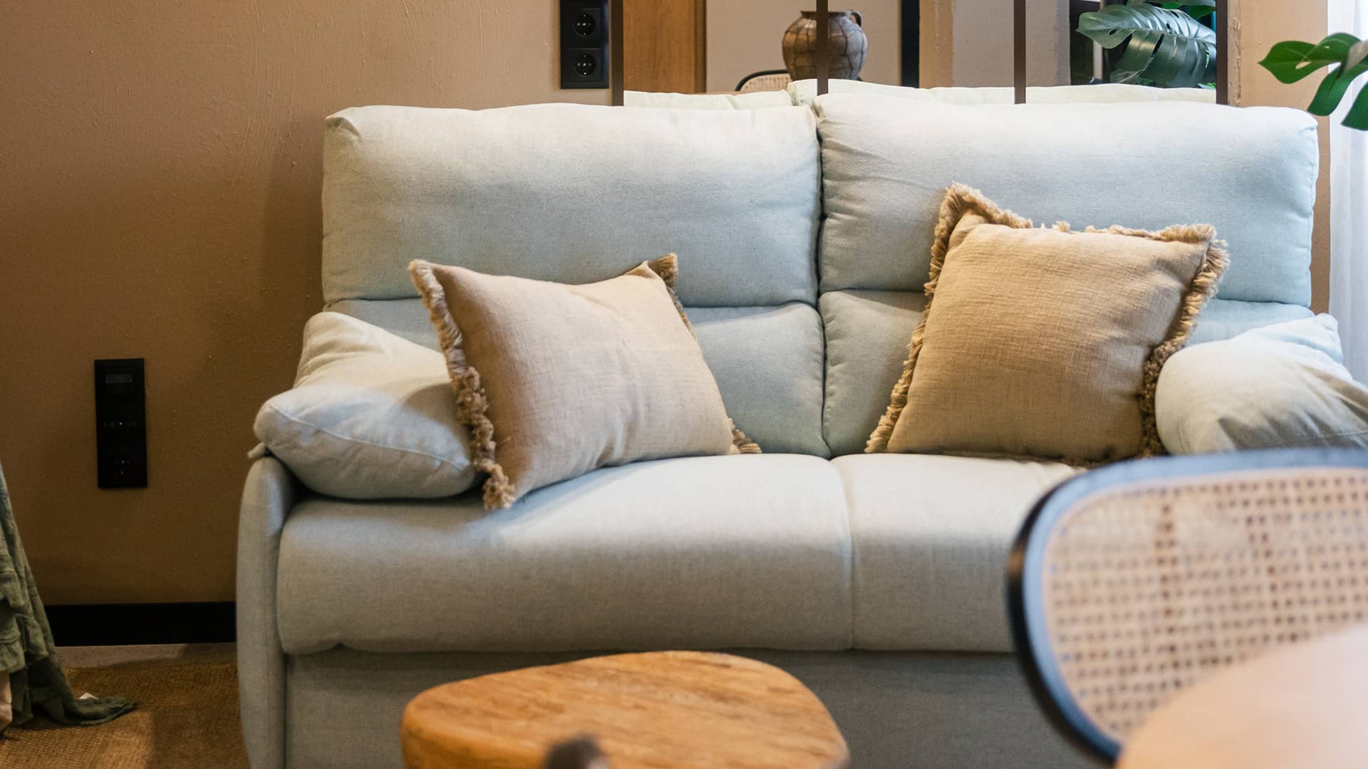 Sofá con cojines de flecos Sofá claro con cojines beige de flecos, mesa baja de madera y silla de rejilla en un apartamento de Grupo CCOMMO