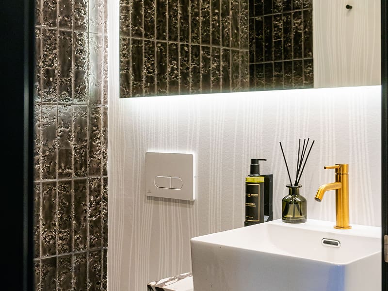 Apartamento Premium con balcón | Baño Lavabo blanco con grifo dorado, jabón y difusor, junto a azulejos verdes retroiluminados