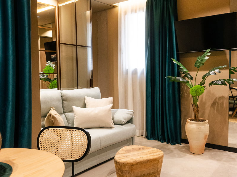 Apartamento Premium | Salón Sofá claro con cojines, mesa de rejilla, taburete de madera, planta y cortinas verdes