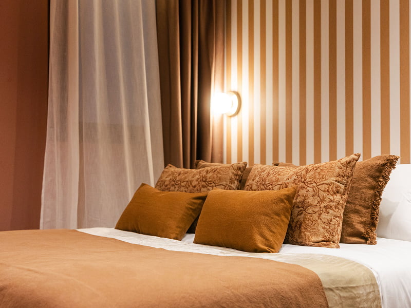 Apartamento Premium | Dormitorio en tonos cálidos Cama con cojines en tonos ocre y terracota frente a una pared de rayas con aplique