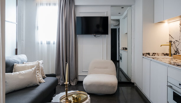 Apartamento para 2 | Salón y cocina Salón con sofá oscuro, butaca blanca y televisión, con cocina blanca de encimera de mármol