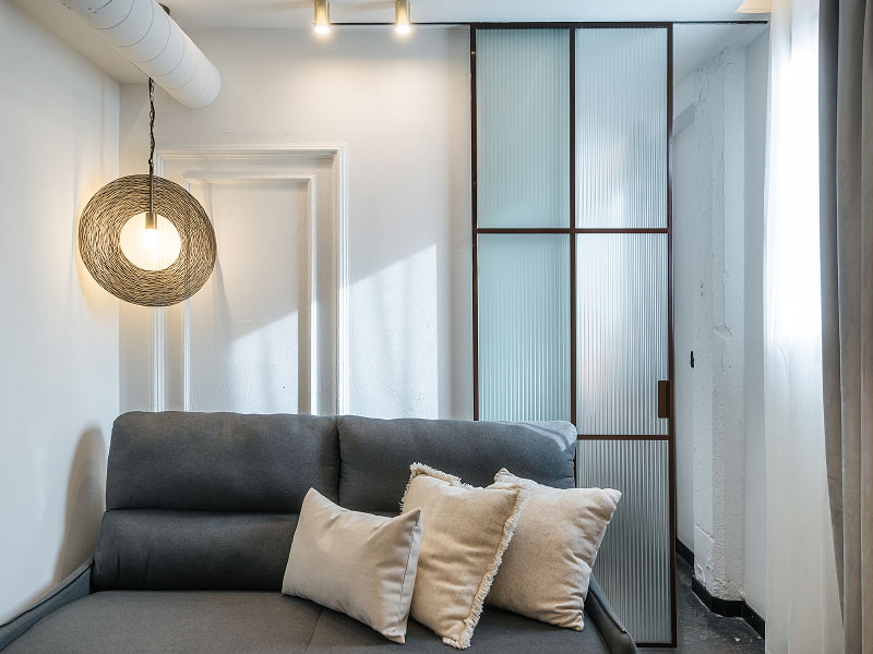 Apartamento para 2 | Sofá Sofá gris con cojines claros bajo una lámpara de mimbre, junto a una mampara de vidrio