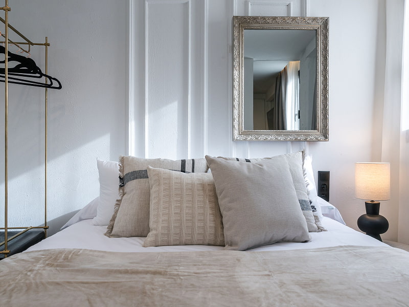 Apartamento para 2 | Dormitorio Cama con cojines grises y beige, espejo de marco plateado y lámpara de mesilla