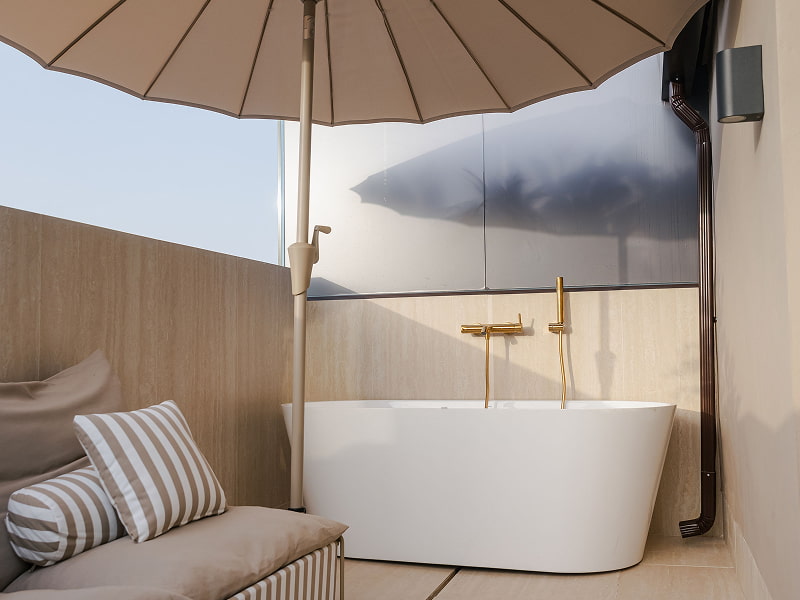 Ático con vistas | Jacuzzi exterior Bañera de hidromasaje exterior con grifería dorada bajo una sombrilla, junto a un sofá con cojines de rayas