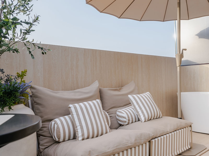 Ático con vistas | Sofá de exterior Sofá de exterior con cojines de rayas beige bajo una sombrilla y rodeado de plantas en la terraza