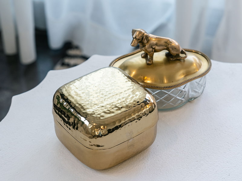 Ático con vistas | Detalles decorativos Cajitas decorativas doradas, una con tapa de cristal y figura de perro, sobre una mesa blanca