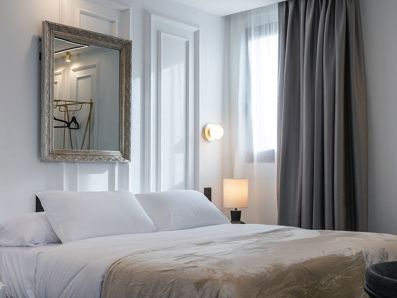 Estudio Premium con balcón | Dormitorio Cama doble con manta beige, espejo de marco plateado, aplique y cortinas grises