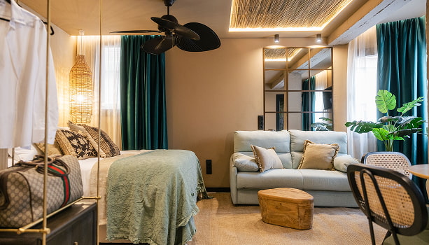 Estudio Premium | Plano general Estudio Premium de Los 36 con cama, sofá, ventilador de techo, cortinas verdes y techo de cañizo iluminado