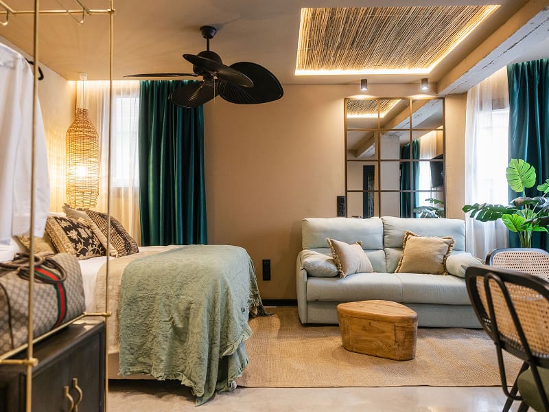 Estudio Premium | Plano general Estudio Premium de Los 36 con cama, sofá, ventilador de techo, cortinas verdes y techo de cañizo iluminado