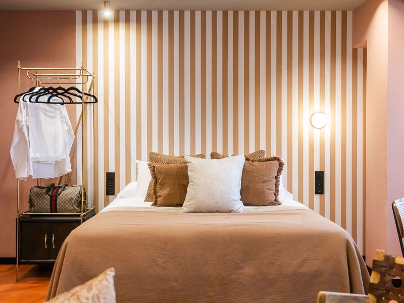 Estudio Premium | Dormitorio en tonos cálidos Cama con cojines en tonos tierra frente a una pared de rayas y perchero con camisas blancas