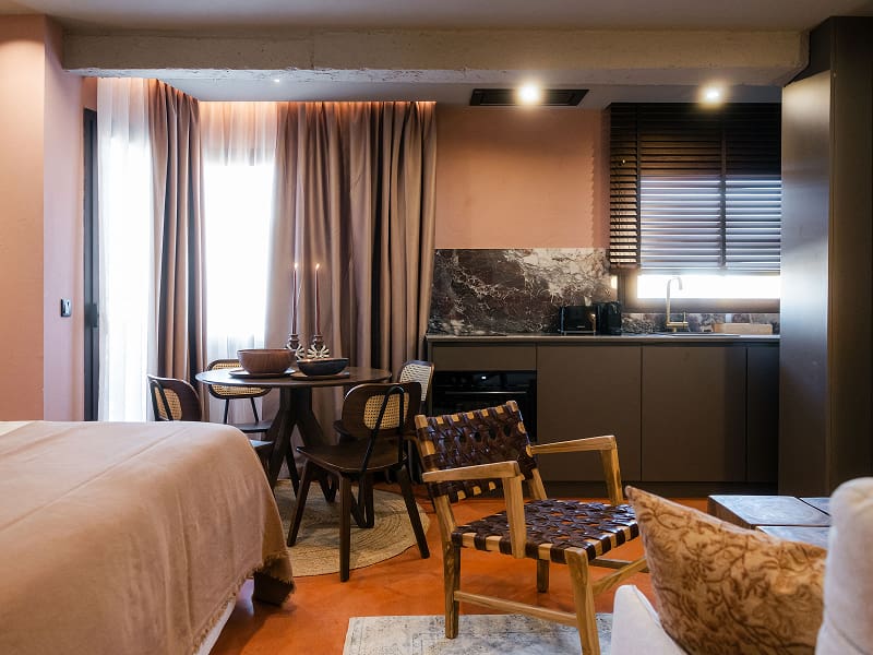 Estudio Premium | Vista general en tonos cálidos Estudio con paredes rosadas, cama, sillón de madera y cuero, comedor y cocina gris al fondo