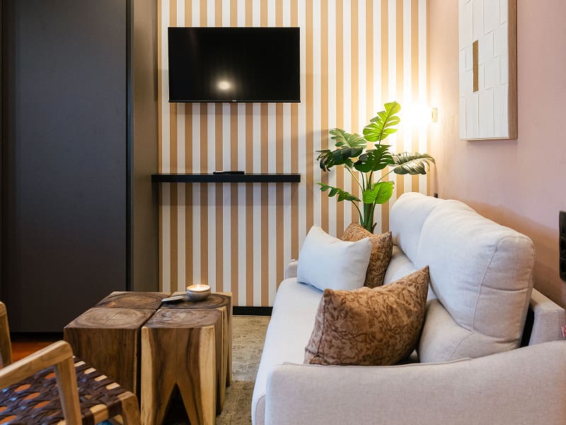 Estudio Premium | Zona de estar Sofá blanco con cojines, mesitas de madera y televisión sobre una pared de rayas