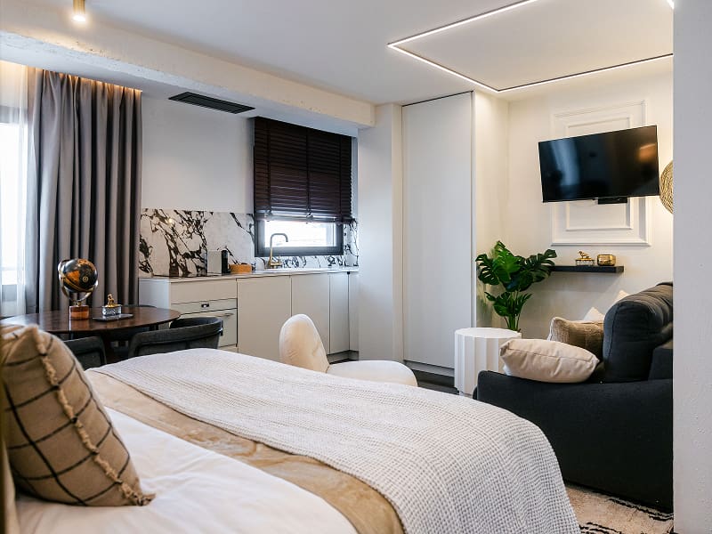 Estudio Premium | Dormitorio y salón Estudio blanco con cama, sofá oscuro, televisión y cocina junto a la ventana