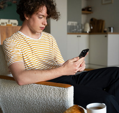 Check-in online desde el móvil Joven con camiseta de rayas usando el móvil sentado en un sillón, con una taza a su lado