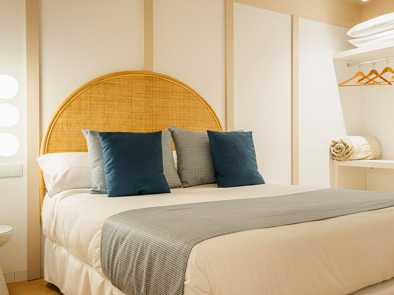 Apartamento 1 habitación | Dormitorio en azul Cama con cabecero de ratán, cojines azul petróleo y manta gris junto a un armario abierto