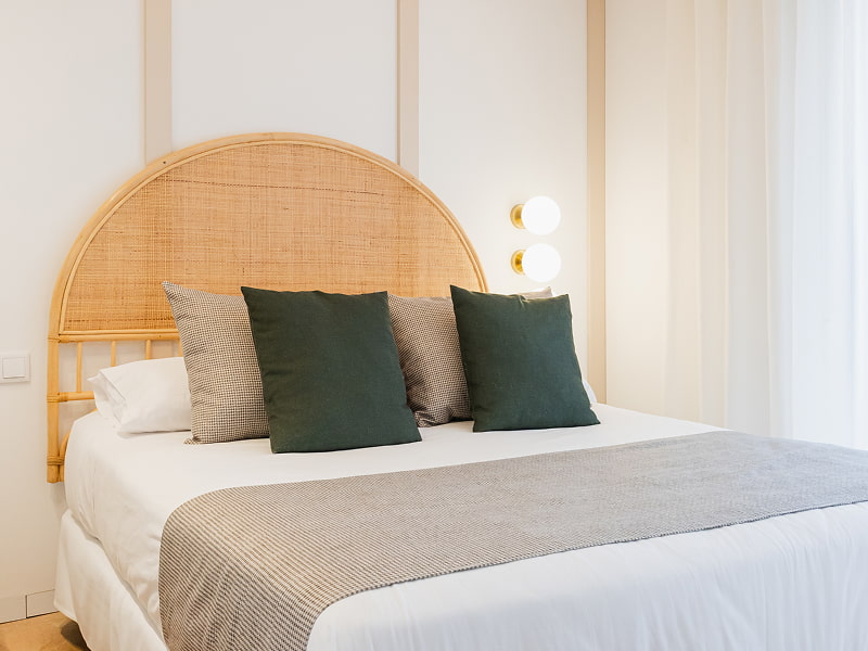 Apartamento 2 habitaciones | Cama doble Cama doble con cabecero de ratán, cojines verde oscuro y manta gris junto a la ventana